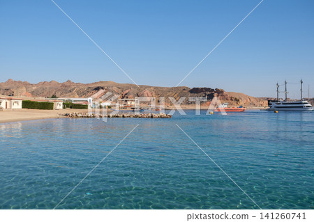 Coastal scene featuring a tranquil beach with clear water, rocky hills in the background, and boats anchored near a small town under a clear sky Coastal scene featuring a tranquil beach with clear water, rocky hills in the background, and boats anchored near a small town under a clear sky 141260741