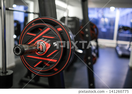 Close-up view of a black and red weight plate on a barbell in a gym, with various exercise machines and equipment visible in the background Close-up view of a black and red weight plate on a barbell in a gym, with various exercise machines and equipment visible in the background 141260775