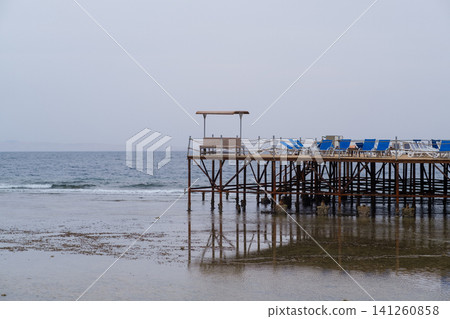 Wooden pier features blue lounge chairs facing the calm sea, with reflections visible in the shallow water under a cloudy sky Wooden pier features blue lounge chairs facing the calm sea, with reflections visible in the shallow water under a cloudy sky 141260858