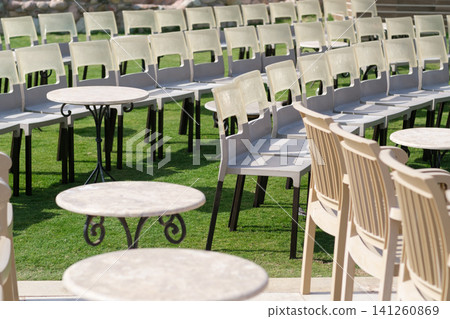 Outdoor setting featuring rows of plastic chairs and small round tables placed on a grassy area, illuminated by natural sunlight Outdoor setting featuring rows of plastic chairs and small round tables placed on a grassy area, illuminated by natural sunlight 141260869