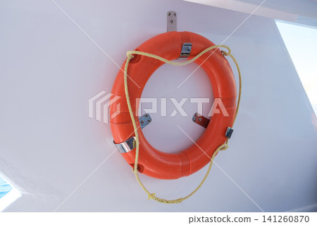 Bright orange life buoy is securely mounted on a white wall, intended for safety on boats and marine vessels in sunny outdoor conditions Bright orange life buoy is securely mounted on a white wall, intended for safety on boats and marine vessels in sunny outdoor conditions 141260870