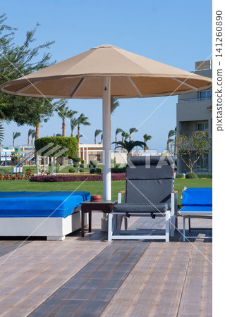 Poolside area features sun loungers with blue cushions, a large umbrella, and a landscaped garden in the background under a clear blue sky Poolside area features sun loungers with blue cushions, a large umbrella, and a landscaped garden in the background under a clear blue sky 141260890