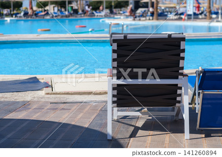 Black and white lounge chair sits next to a clear swimming pool, with additional sun loungers and umbrellas visible in the background under bright sunlight Black and white lounge chair sits next to a clear swimming pool, with additional sun loungers and umbrellas visible in the background under bright sunlight 141260894