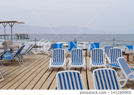 Beachside deck features striped lounge chairs on wooden planks, with calm ocean waters and distant mountains visible in the background Beachside deck features striped lounge chairs on wooden planks, with calm ocean waters and distant mountains visible in the background 141260909