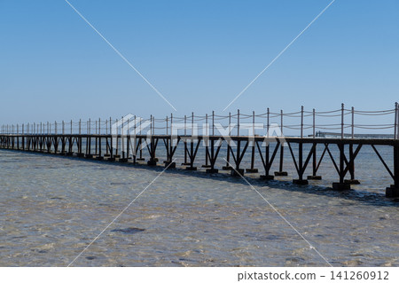 Wooden pier stretches over clear water, with a bright blue sky above and a distant shoreline visible, creating a tranquil coastal scene Wooden pier stretches over clear water, with a bright blue sky above and a distant shoreline visible, creating a tranquil coastal scene 141260912