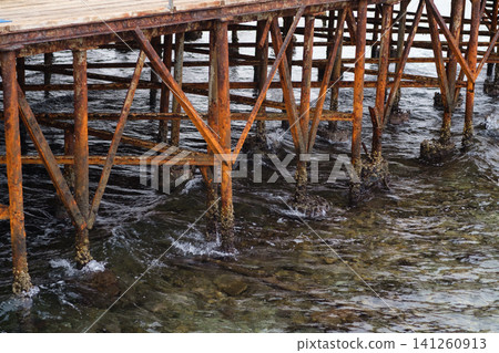 Weathered wooden pier structure features rusted supports above clear water, with visible barnacles on the pilings in a coastal setting Weathered wooden pier structure features rusted supports above clear water, with visible barnacles on the pilings in a coastal setting 141260913