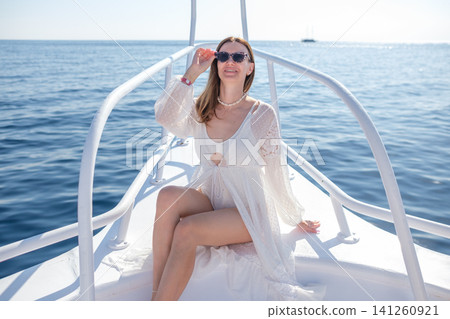 Woman in white beach cover-up relaxes on the bow of a boat, wearing sunglasses, with calm ocean waters and a clear sky in the background Woman in white beach cover-up relaxes on the bow of a boat, wearing sunglasses, with calm ocean waters and a clear sky in the background 141260921