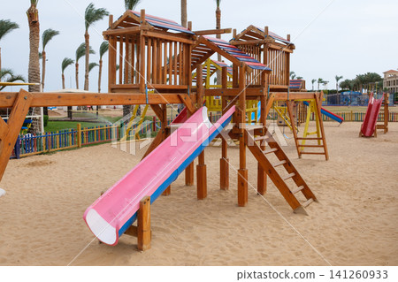 Wooden playground equipment features a pink slide and climbing elements in a sandy area, with palm trees and colorful fencing in the background Wooden playground equipment features a pink slide and climbing elements in a sandy area, with palm trees and colorful fencing in the background 141260933