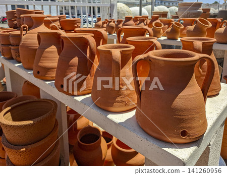 A display has many terracotta water pitchers and ceramic vessels on outdoor shelves. The setting is ideal for home decoration and gardening tasks in a sunny location. A display has many terracotta water pitchers and ceramic vessels on outdoor shelves. The setting is ideal for home decoration and gardening tasks in a sunny location. 141260956