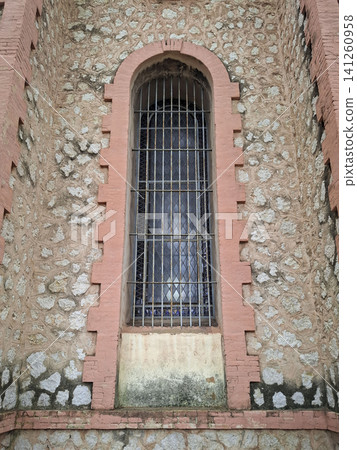 A tall, arched stained glass window sits in a stone wall of an abandoned church in Spain. The window is behind a rusted iron security grate. The church shows signs of age and neglect. A tall, arched stained glass window sits in a stone wall of an abandoned church in Spain. The window is behind a rusted iron security grate. The church shows signs of age and neglect. 141260958