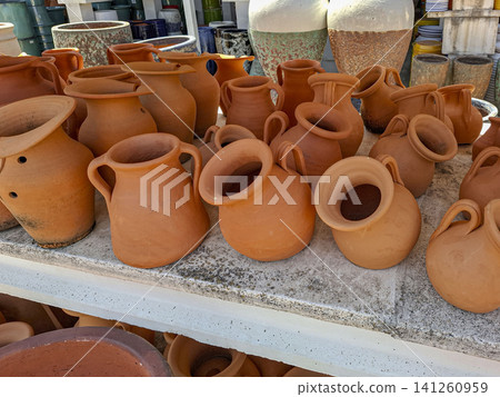 Handmade terracotta clay pots and ceramic jugs are on display at a rustic market. The items represent traditional craft and add to the Mediterranean decor. Shoppers explore the unique creations. 141260959
