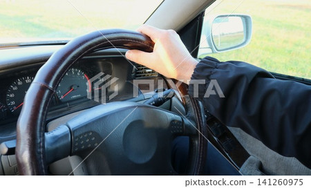 view from inside the car: a driver in a dark windbreaker holds the top of the steering wheel. The speedometer, tachometer, and air vent are visible, and the roadside is reflected in the outside mirror view from inside the car: a driver in a dark windbreaker holds the top of the steering wheel. The speedometer, tachometer, and air vent are visible, and the roadside is reflected in the outside mirror 141260975