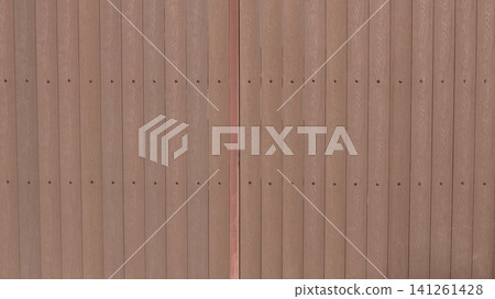 A brown fence made of vertical planks with a textured wood grain. The planks are screwed to the metal frame with even rows of screws, and a vertical joint is visible in the center of the frame. A brown fence made of vertical planks with a textured wood grain. The planks are screwed to the metal frame with even rows of screws, and a vertical joint is visible in the center of the frame. 141261428