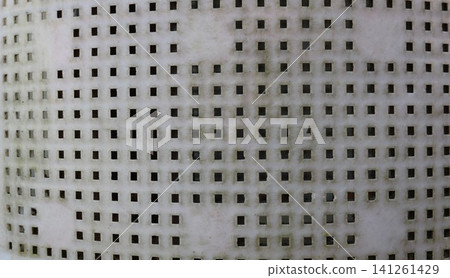 A plastic surface with a regular grid of square holes. The material shows signs of wear, gray dust, and a light dark coating inside some of the cells. An abstract, dirty, perforated background. A plastic surface with a regular grid of square holes. The material shows signs of wear, gray dust, and a light dark coating inside some of the cells. An abstract, dirty, perforated background. 141261429