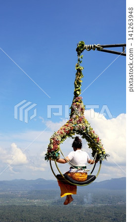 Balinese Woman seated on ornate floral swing above lush valley during Balinese temple celebration, wearing white lace top and traditional sarong, with bright blue sky ,floral swing Balinese Woman seated on ornate floral swing above lush valley during Balinese temple celebration, wearing white lace top and traditional sarong, with bright blue sky ,floral swing 141263948