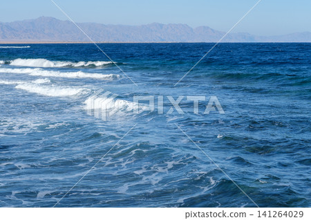 Ocean waves crash softly onto a sandy beach, with distant mountains and a clear blue sky creating a serene coastal landscape Ocean waves crash softly onto a sandy beach, with distant mountains and a clear blue sky creating a serene coastal landscape 141264029