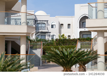 Modern residential buildings feature balconies and staircases, surrounded by palm trees in a landscaped courtyard area with clear blue skies Modern residential buildings feature balconies and staircases, surrounded by palm trees in a landscaped courtyard area with clear blue skies 141264030