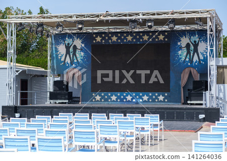 Outdoor stage features a large screen and decorative backdrop, with rows of white chairs arranged for an audience in a sunny setting Outdoor stage features a large screen and decorative backdrop, with rows of white chairs arranged for an audience in a sunny setting 141264036
