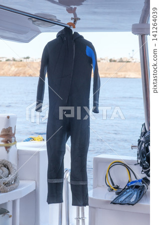 Black wetsuit suspended on a boat, accompanied by diving gear and an ocean view, highlighting water sports equipment and marine scenery Black wetsuit suspended on a boat, accompanied by diving gear and an ocean view, highlighting water sports equipment and marine scenery 141264039