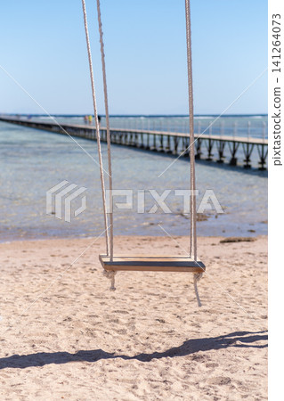 Wooden swing suspended by ropes on a sandy beach, with a long pier leading into the calm sea and a clear blue sky above 141264073