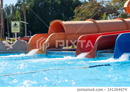 Colorful water slides are positioned above a swimming pool with clear blue water, surrounded by greenery and a sunny atmosphere Colorful water slides are positioned above a swimming pool with clear blue water, surrounded by greenery and a sunny atmosphere 141264074