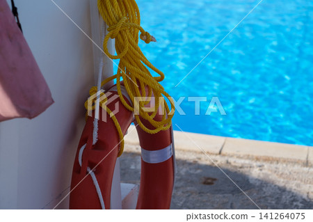 Red lifebuoy secured with yellow rope on a wall beside a swimming pool featuring clear blue water in the background Red lifebuoy secured with yellow rope on a wall beside a swimming pool featuring clear blue water in the background 141264075