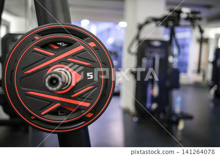 Close-up view of a black and red weight plate on a barbell in a contemporary gym, with various exercise machines in the background Close-up view of a black and red weight plate on a barbell in a contemporary gym, with various exercise machines in the background 141264078