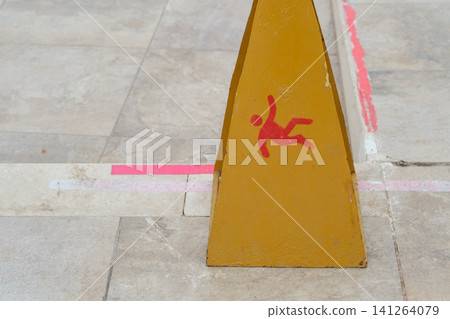 Yellow caution sign displays a red figure falling, placed on a tiled floor with visible markings, warning of a slip hazard in a public space Yellow caution sign displays a red figure falling, placed on a tiled floor with visible markings, warning of a slip hazard in a public space 141264079