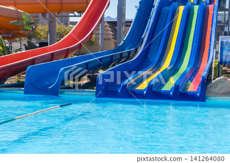 Colorful water slides in red, blue, and yellow positioned next to a clear swimming pool with palm trees visible in the background Colorful water slides in red, blue, and yellow positioned next to a clear swimming pool with palm trees visible in the background 141264080