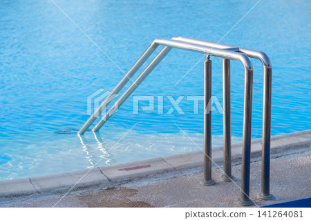 Stainless steel ladder descends into a clear blue swimming pool, featuring a tiled edge and reflections of sunlight on the water surface Stainless steel ladder descends into a clear blue swimming pool, featuring a tiled edge and reflections of sunlight on the water surface 141264081