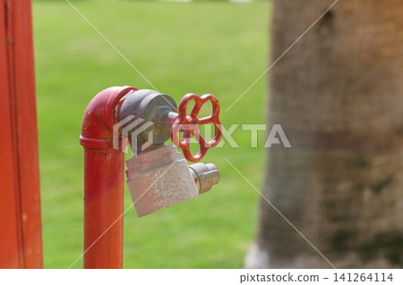 Red fire hydrant valve features a flower-shaped handle, situated in front of a lush green lawn, with a blurred tree trunk in the background Red fire hydrant valve features a flower-shaped handle, situated in front of a lush green lawn, with a blurred tree trunk in the background 141264114