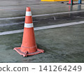 An orange plastic safety cone with white stripes placed on a grey concrete floor inside a building or parking area. Concept for caution, workplace safety, traffic control, and warning. 141264249