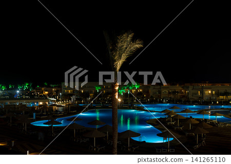 Night view of a brightly lit swimming pool surrounded by palm trees and lounge chairs at a resort, creating a tranquil atmosphere Night view of a brightly lit swimming pool surrounded by palm trees and lounge chairs at a resort, creating a tranquil atmosphere 141265110