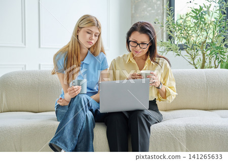 Mother and teenage daughter looking at laptop screen together on sofa 141265633