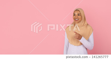 Cheerful young Black Muslim woman in beige hijab pointing toward copy space Cheerful young Black Muslim woman in beige hijab pointing toward copy space 141265777