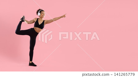 Flexible young Black fitness woman wearing headphones performing standing leg stretch Flexible young Black fitness woman wearing headphones performing standing leg stretch 141265779