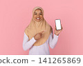 Smiling Black Muslim woman in beige hijab pointing to blank smartphone screen 141265869