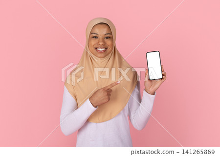 Smiling Black Muslim woman in beige hijab pointing to blank smartphone screen Smiling Black Muslim woman in beige hijab pointing to blank smartphone screen 141265869