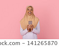 Cheerful young Black Muslim woman in beige hijab holding smartphone in hands 141265870