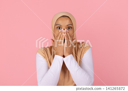 Surprised young Black Muslim woman in hijab covering mouth with both hands Surprised young Black Muslim woman in hijab covering mouth with both hands 141265872