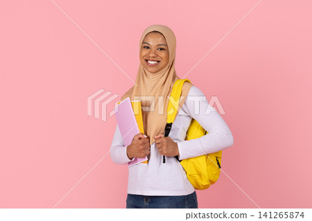 Black Muslim female student in hijab holding colorful notebooks and yellow backpack Black Muslim female student in hijab holding colorful notebooks and yellow backpack 141265874