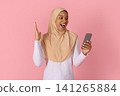 Excited Black Muslim woman in hijab reacting with surprise while holding smartphone 141265884