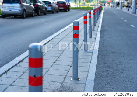 A Close-Up View of Street Bollards with Red Reflective Bands, Providing Safety and Guidance Along a Sidewalk Next to a Busy Vehicle Road 141268082