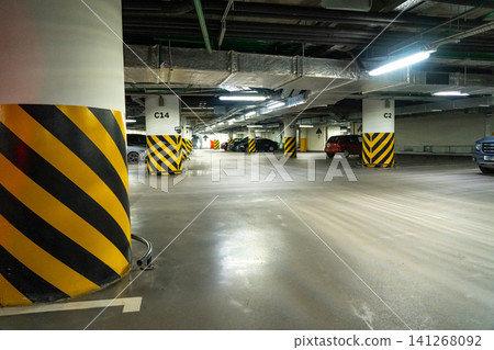 Dimly Lit Parking Garage with Yellow and Black Warning Stripes on Columns and Parked Vehicles Amidst an Open Space Dimly Lit Parking Garage with Yellow and Black Warning Stripes on Columns and Parked Vehicles Amidst an Open Space 141268092