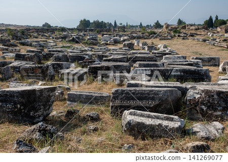 Visitors wander through the historic site of Pamukkale, observing ancient stone remains scattered across the grassy landscape on a sunny day. Visitors wander through the historic site of Pamukkale, observing ancient stone remains scattered across the grassy landscape on a sunny day. 141269077