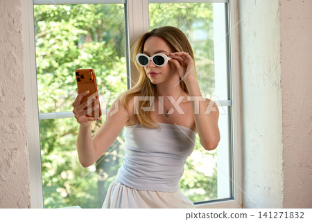 Woman trying white sunglasses while checking appearance with smartphone at home. 141271832