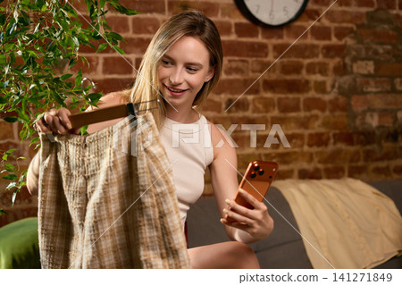 Smiling woman choosing plaid skirt while browsing fashion products on smartphone. Smiling woman choosing plaid skirt while browsing fashion products on smartphone. 141271849