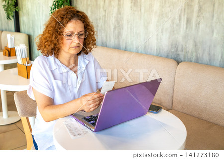 Mature woman checking receipt and managing finances on a laptop in a cozy cafe 141273180