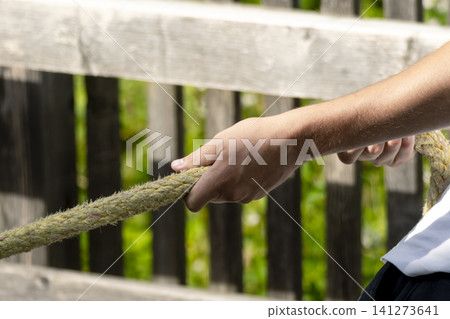 Person pulling a yellow braided rope against a wooden fence and foliage, illustrating the boat docking and mooring process. Person pulling a yellow braided rope against a wooden fence and foliage, illustrating the boat docking and mooring process. 141273641
