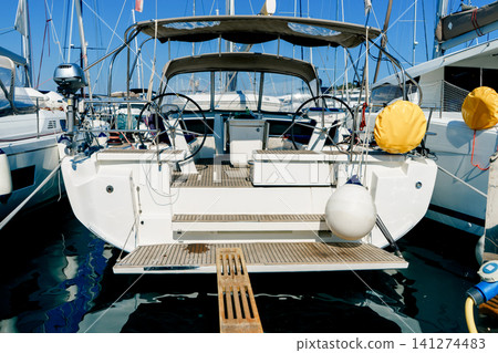 Rear view of a modern sailing yacht moored at the marina, featuring twin helm wheels, teak deck and protective fenders on a sunny day Rear view of a modern sailing yacht moored at the marina, featuring twin helm wheels, teak deck and protective fenders on a sunny day 141274483
